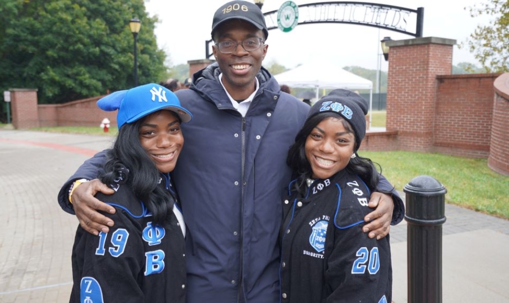 An African American man with a baseball hat and glasses poses with two African American women wearing sorority-oriented jackets