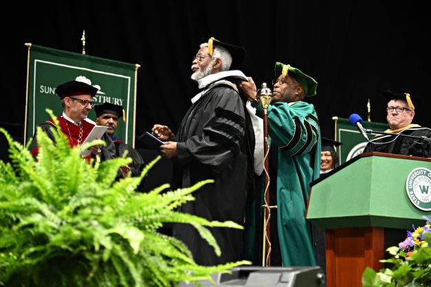 A Black man in green regalia drapes a hood over the head of a Black man in black regalia