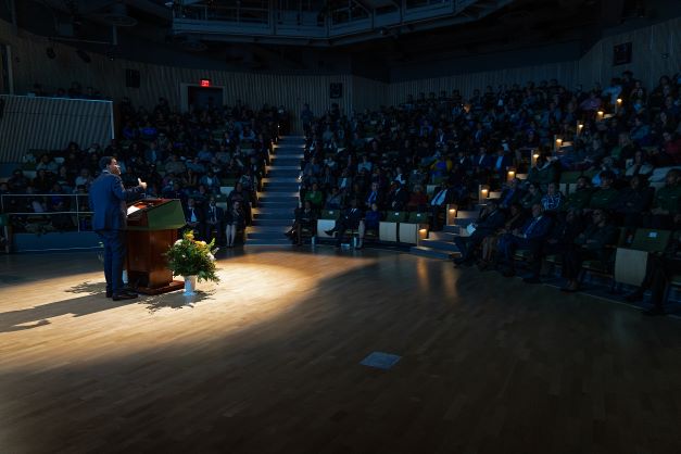 African American man stands behind podium in a spotlight addressing a packed house