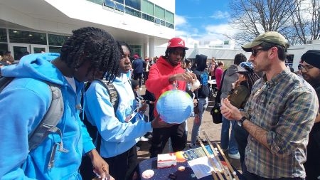 Three young males listen as a professor points to a solar system model