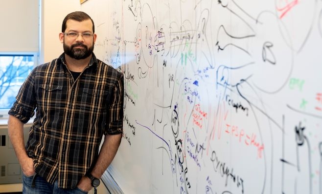 Man in plaid shirt in a classroom leaning against a white board 