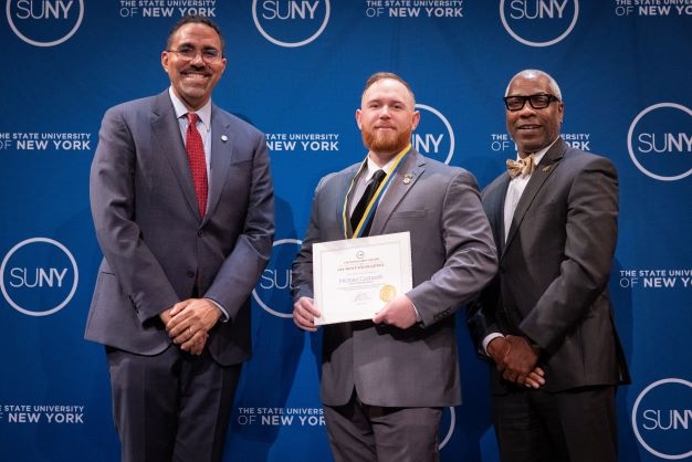 Two adult black men stand on either side of a young white man holding an award certificate