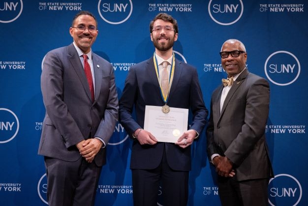 Two adult black men stand on either side of a young white man holding an award certificate