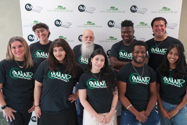 A diverse group of 10 people wearing black tee shirts standing in front of a step and repeat featuring the college logo and radio station logo