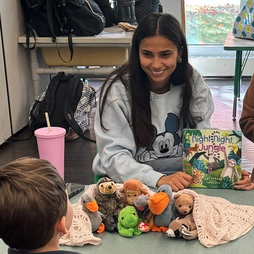 A young woman reading a children's book with an array of stuffed animals on the table in front of her