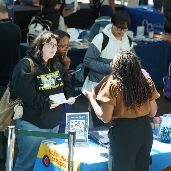 Woman in black sweatshirt and green pants speaks with another woman with black pants and a brown blouse during a networking event