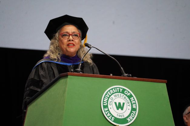 Older Latina woman in black regalia speaks from a podium featuring the green and white seal of SUNY Old Westbury