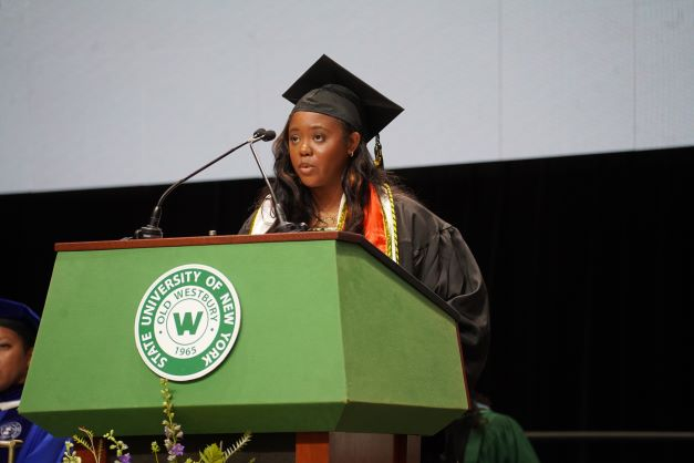 Young African American woman in black regalia speaks from a podium decorate with a green and white seal for SUNY Old Westbury