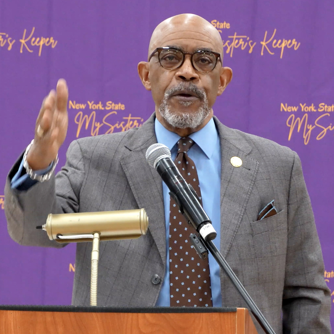 Bald African American man with glasses dressed in a suit speaks from behind a podium