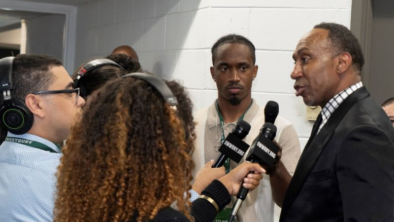 Black man in suit speaks into multiple hand held microphones in an interview scrum