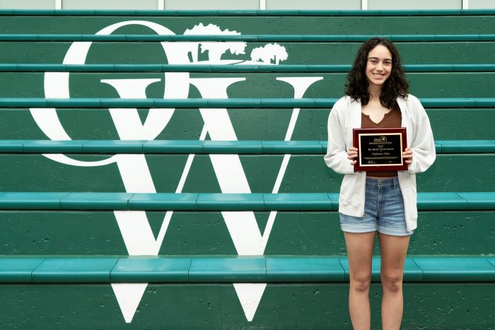 Young woman wearing shirt and long shirt stands in front of green steps holding an award plaque