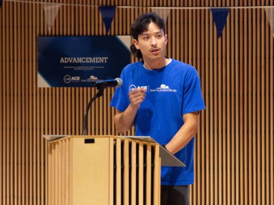 Hispanic male students in blue ACE tee shirts speaks from a podium