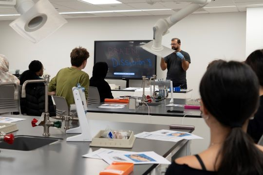 Professor in black shirt lectures to an audience of students as he stands next to a screen that reads Brain Dissection