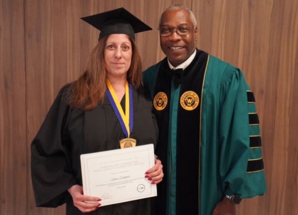 A woman in black regalia accepts an award certificate from a man in green regalia