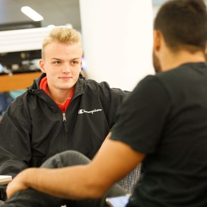 White man in black jacket seated while speaking with another man