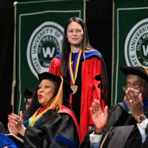 Asian American woman in red regalia stands on stage during commencement
