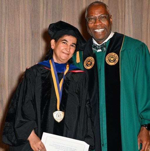 South Asian woman dressed in black regalia accepts congratulations from an African American man in green regalia