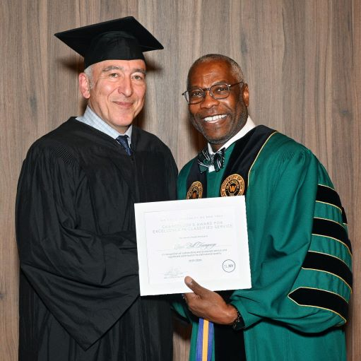 Caucasian man in black regalia accepts a certificate from an African American man in green regalia