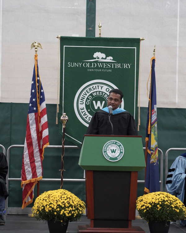 Man in ceremonial regalia speaking into microphone at podium. SUNY Old Westbury seal behind him alongside country and state flags.