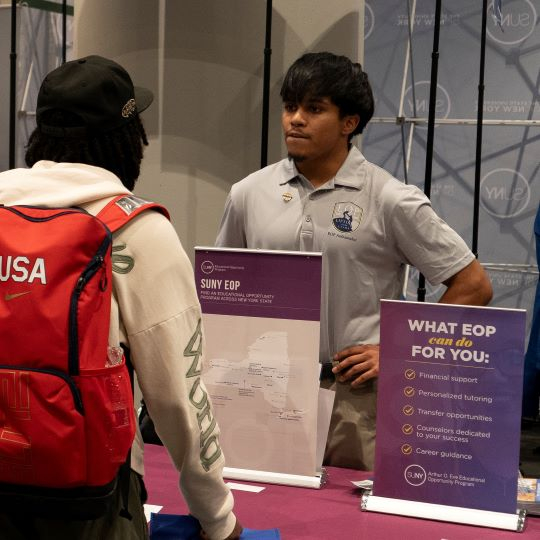 Hispanic male wearing short-sleeved gray shirt speaks with another student over a table 