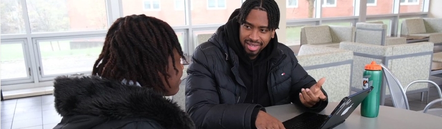 Male student in black jacket seated while speaking with another student