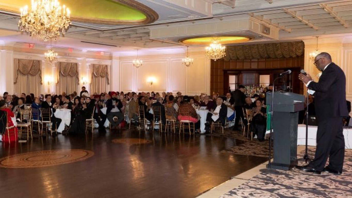 Ballroom filled with people in formal attire while African American man in tuxedo speaks from a podium