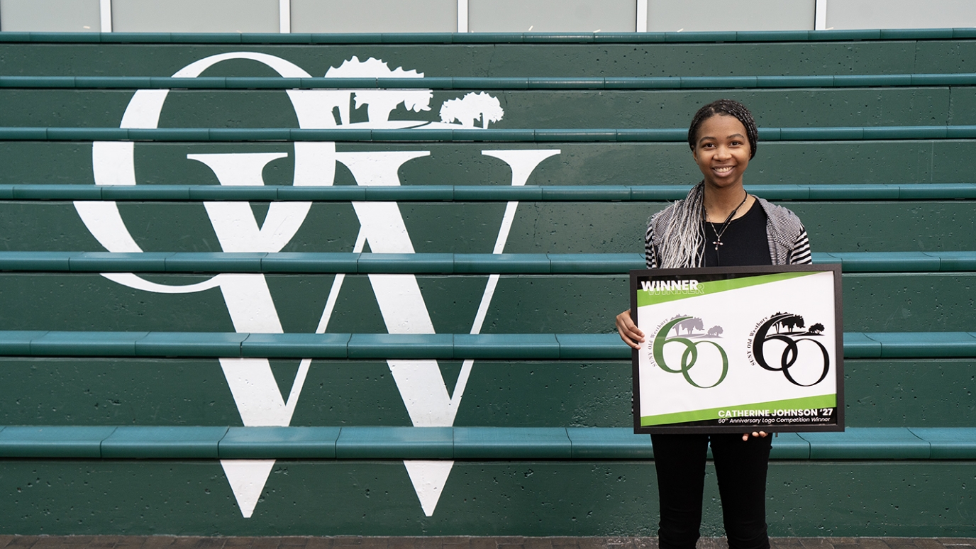 African American young woman stands before a flight of green steps with letters O and W in white while holding a framed print of a logo featuring the number 60  