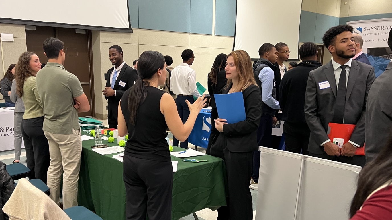 Students and professionals smiling and talking on two sides of a table. Other students with backs towards camera in background.