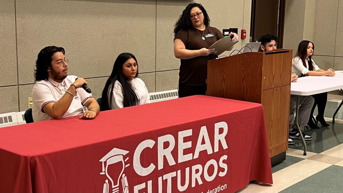 Four students seated at tables with microphones before a diverse group of students