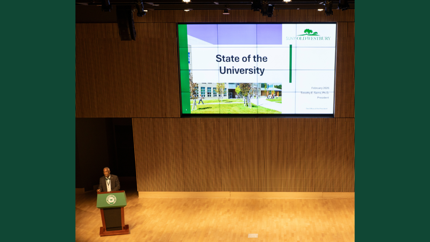 African American man speaking from a podium with a large screen with text reading State of the University 2026