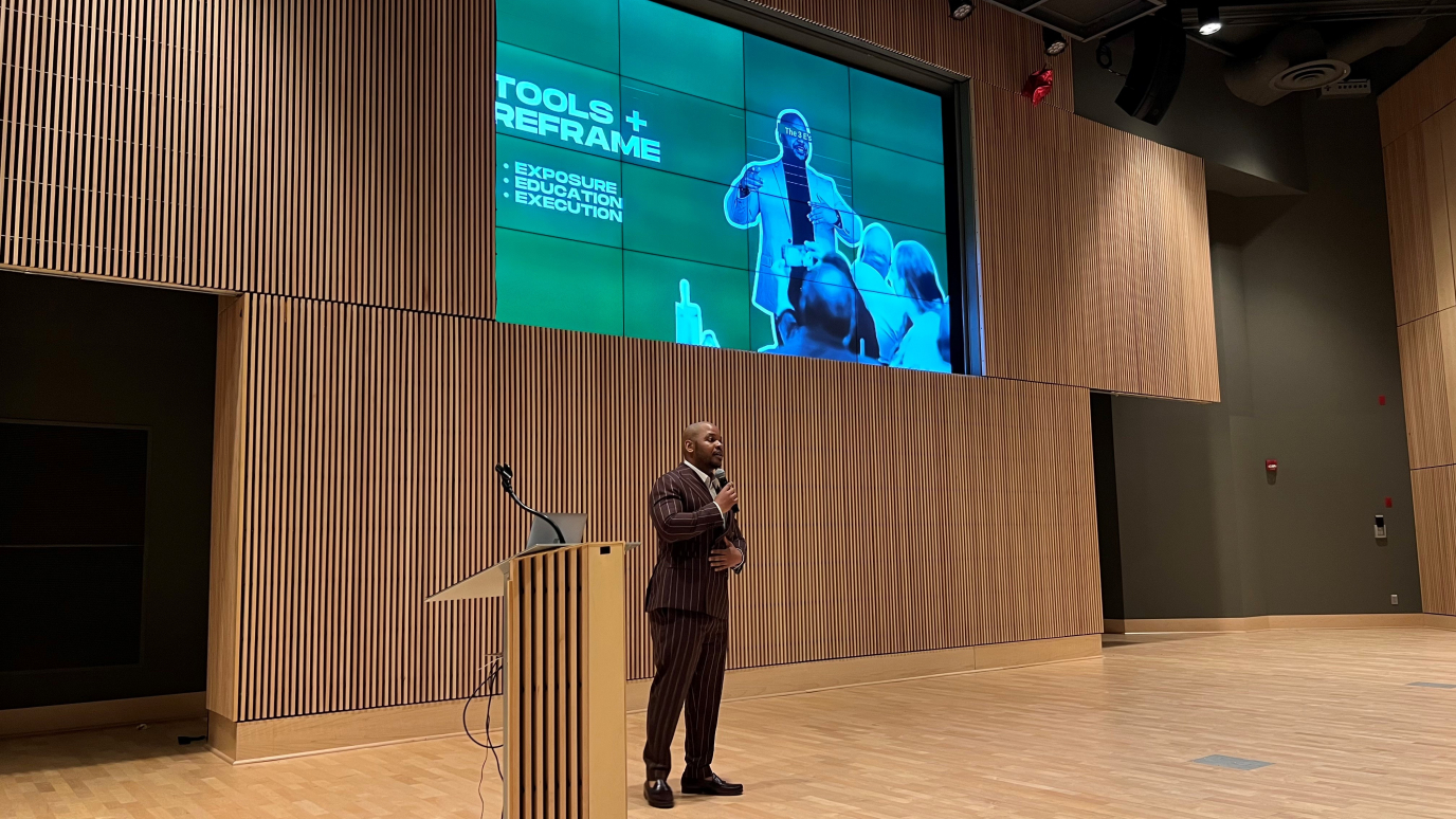 African American man in suit speaks from a stage with a large presentation screen behind him
