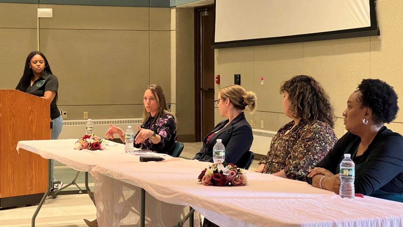 An African American young woman at podium with four other professional women seated at a table