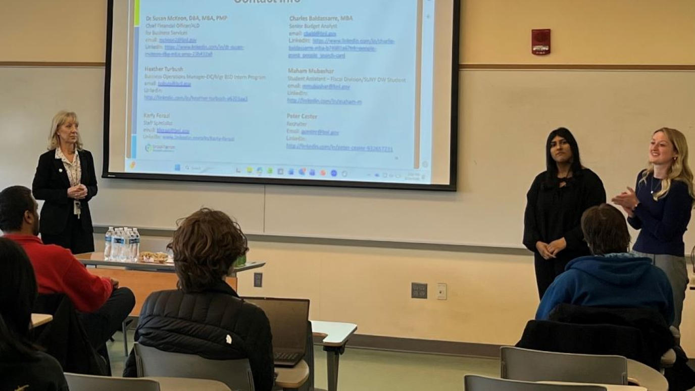 Three women in professional attire speak before a classroom of students with a presentation showing on the room's lightboard