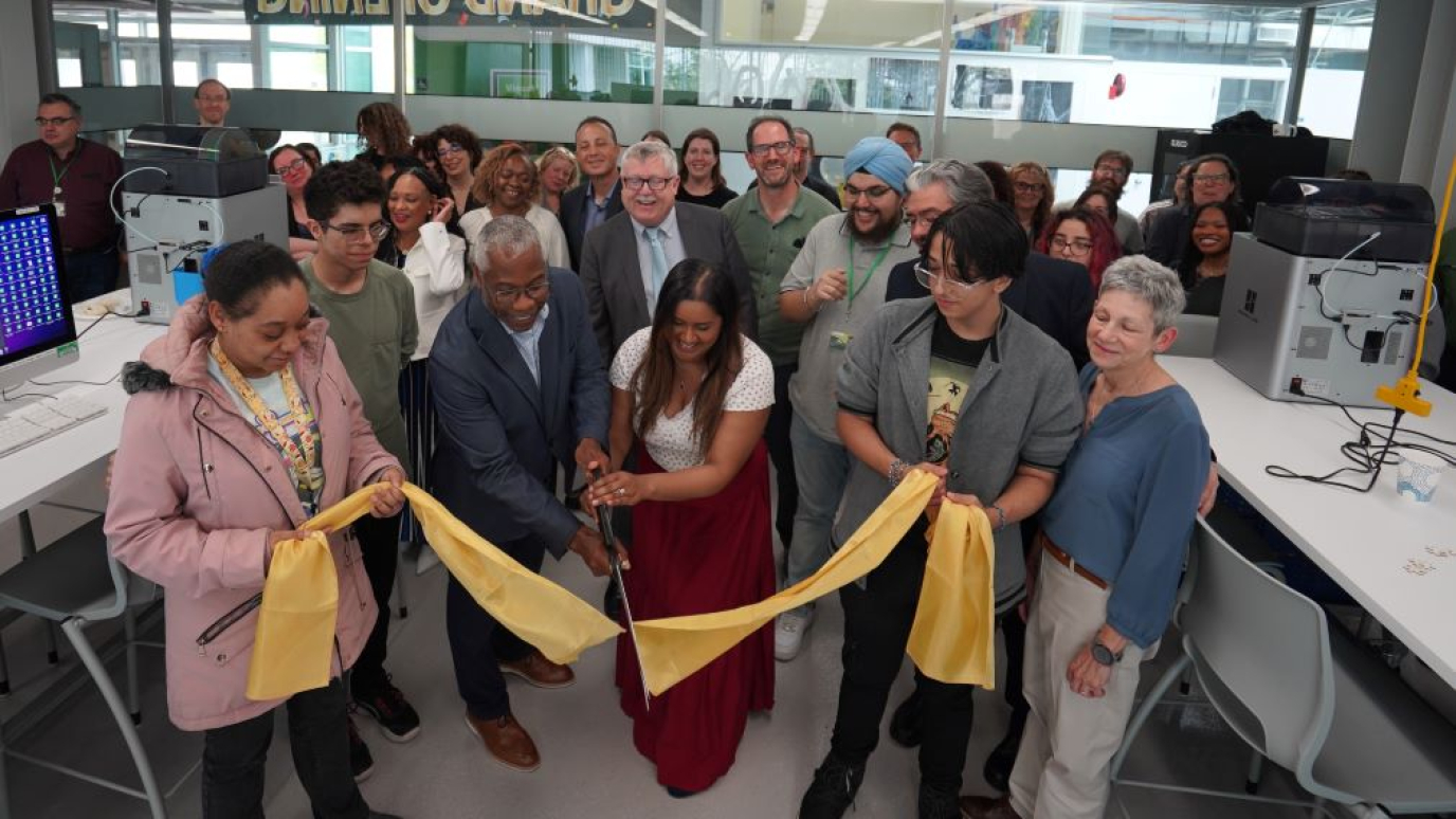 An African American man and a South Asian young woman hold a large pair of scissors above a recently cut ribbon as a crowd of 40 students and professors applaud