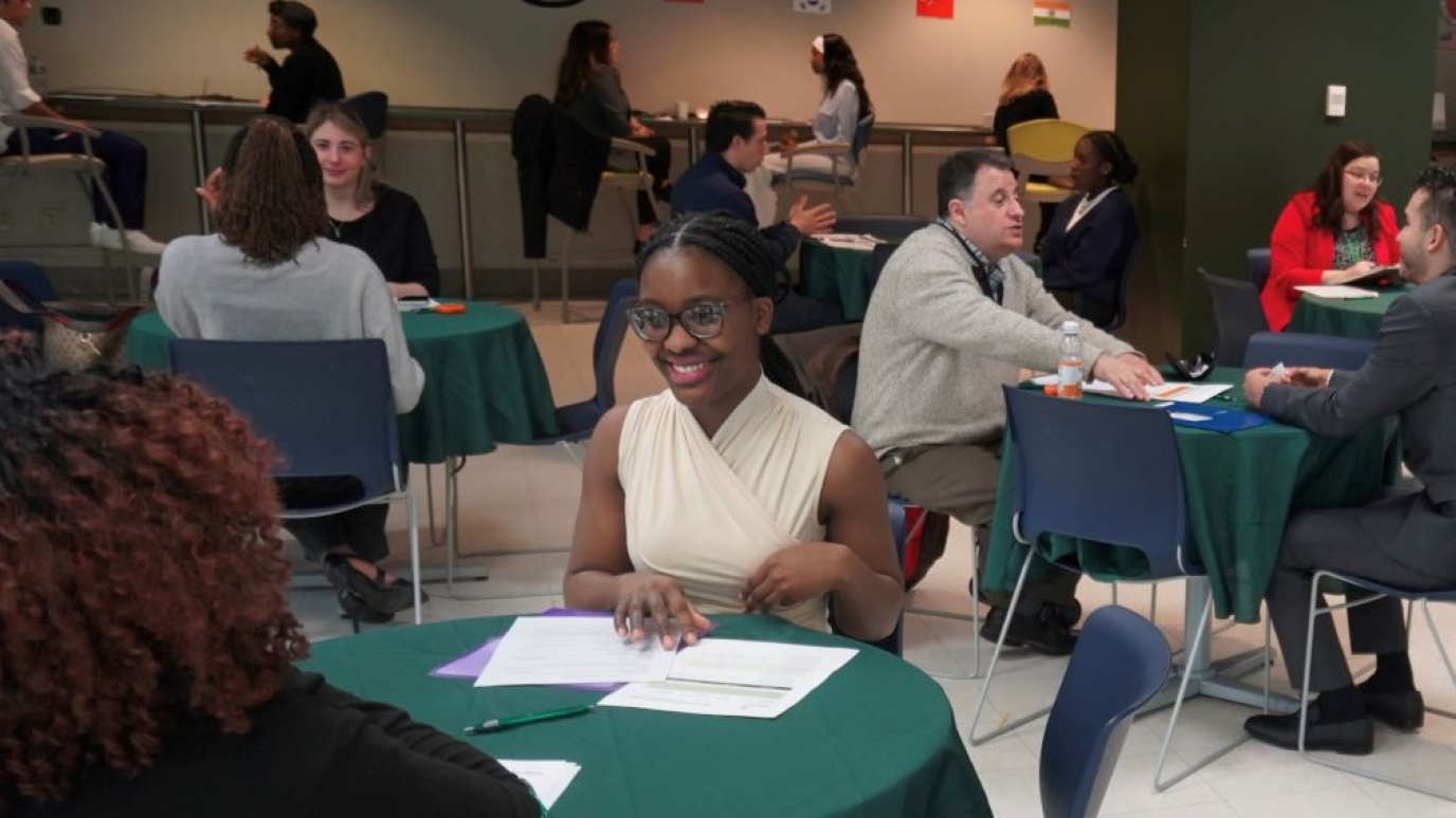Young African American woman speaking at a table in a one-one-conversation surrounded by other tables with pairs of people in conversation