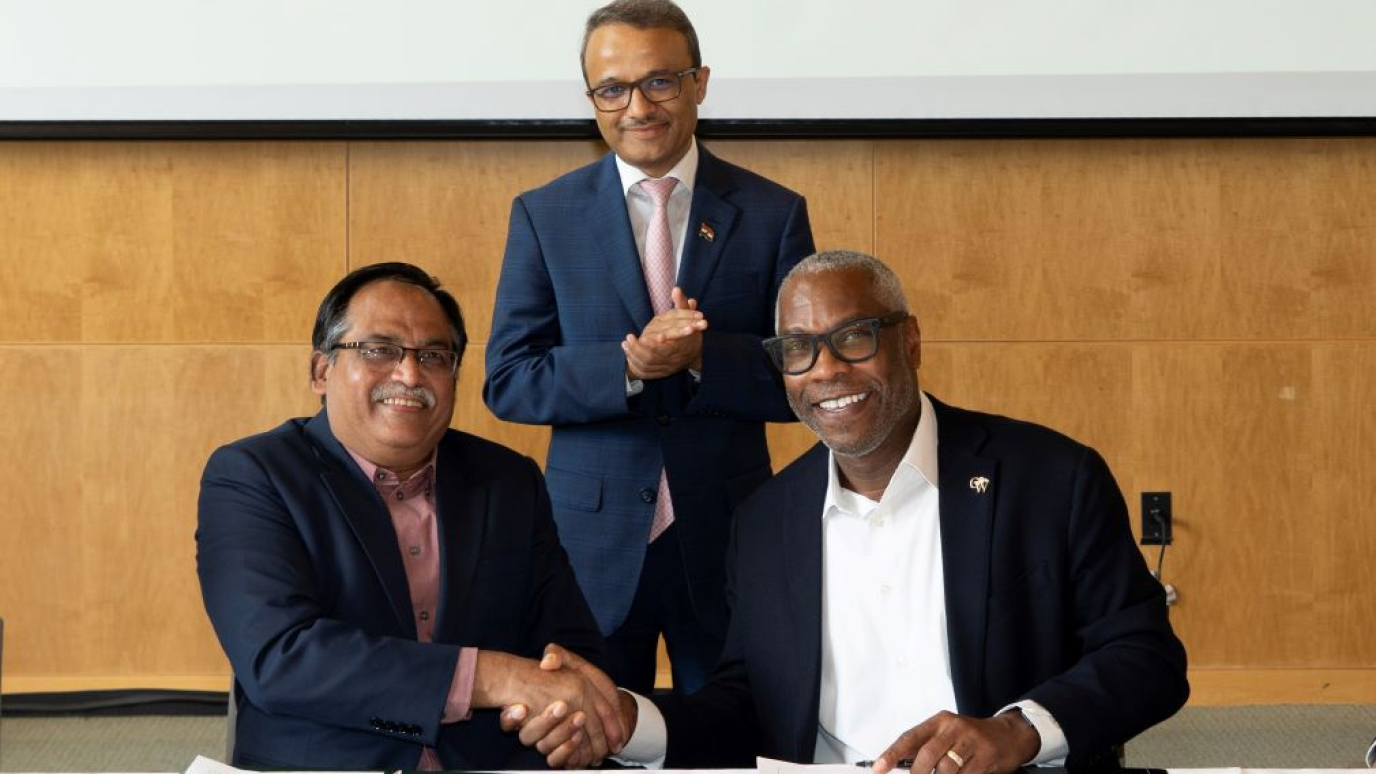 A man of South Asian descent and an African American man shake hands while smiling as another man of South Asian descent looks on