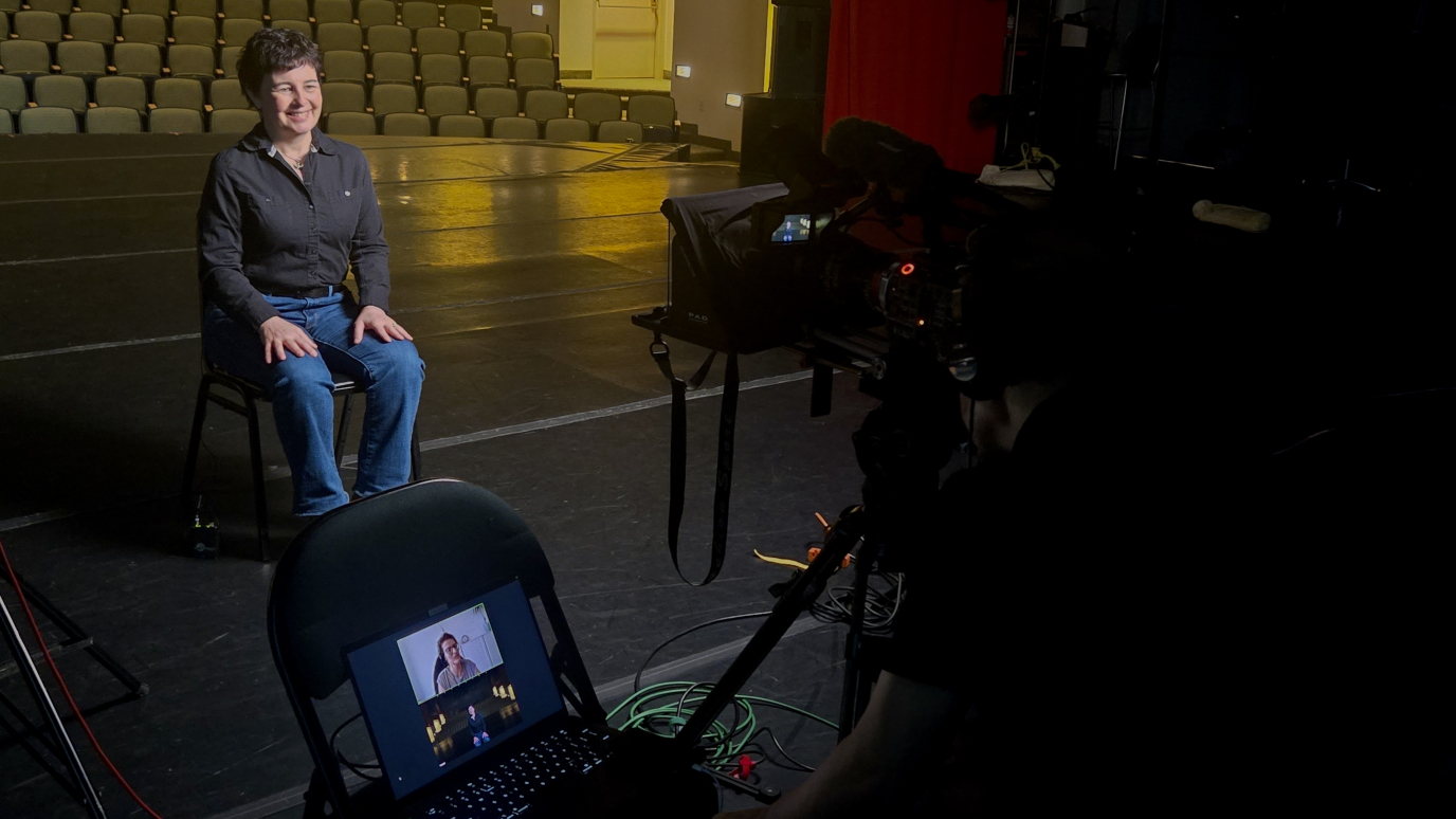 Professor sitting and smiling on chair on Maguire Theater stage while looking towards a camera. Video conference streaming on laptop on chair.