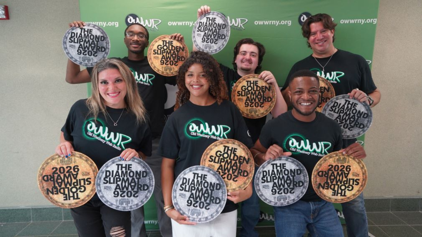 A diverse group of six people wearing black tee shirts standing in front of a step and repeat featuring the radio station logo