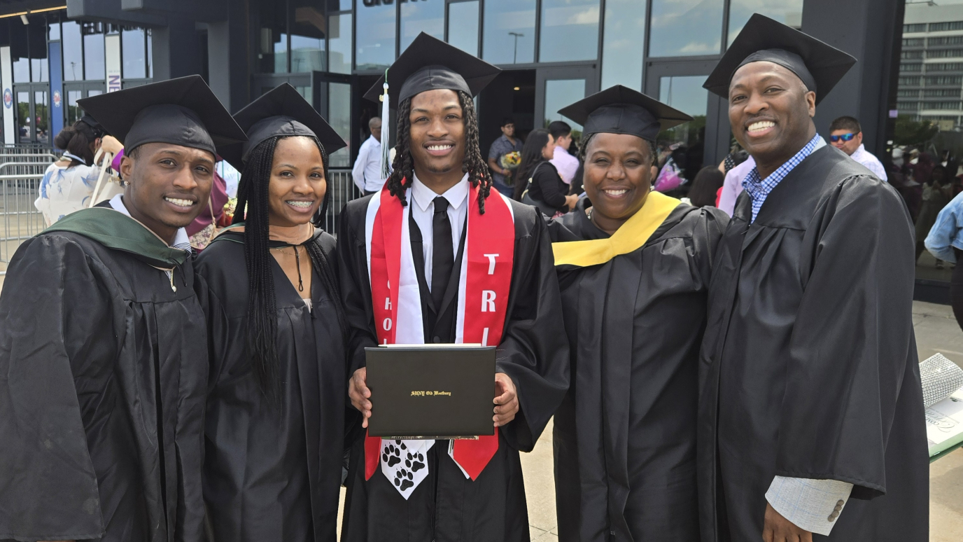 Group of five wearing Commencement regalia standing and smiling together as young man in the middle holds a diploma cover.