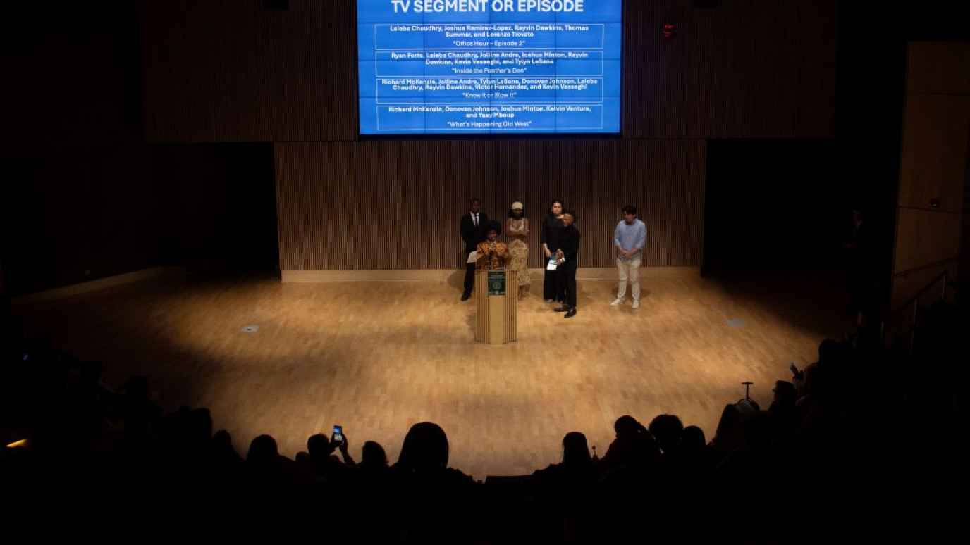 Five students on a brightly lit stage before a large audience cast in shadow