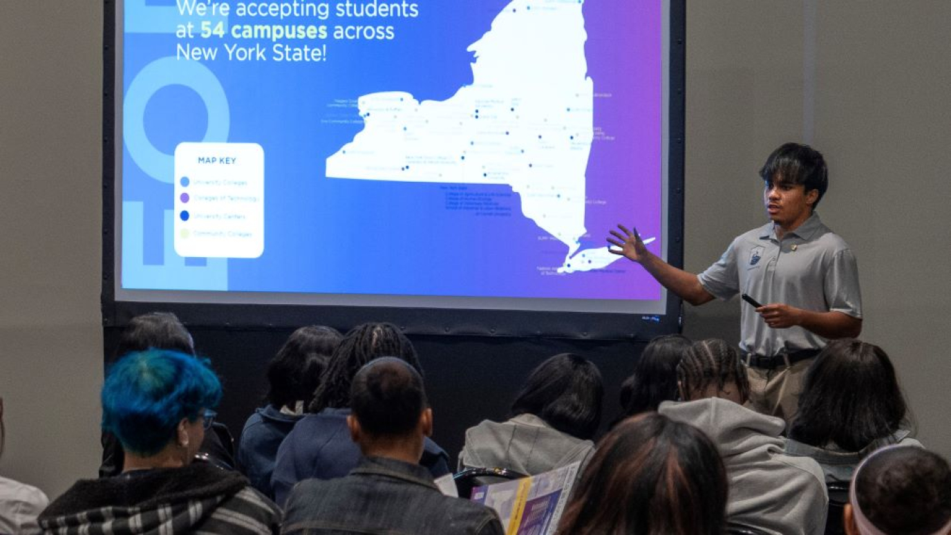 Hispanic male wearing short-sleeved gray shirt presents before a screen showing an outline of New York State.