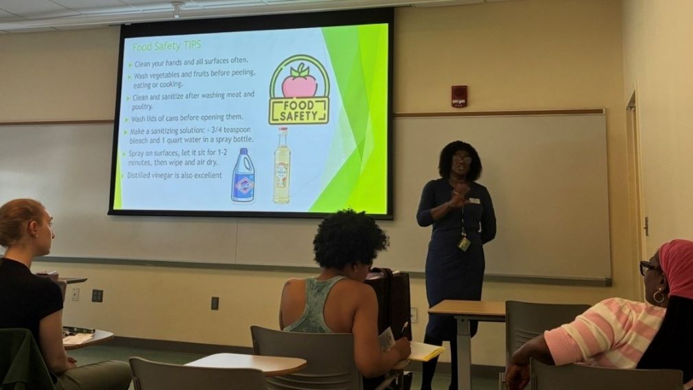 African American woman in professional dress lectures to a group of women while standing in front of a screen that shows a slide on food safety