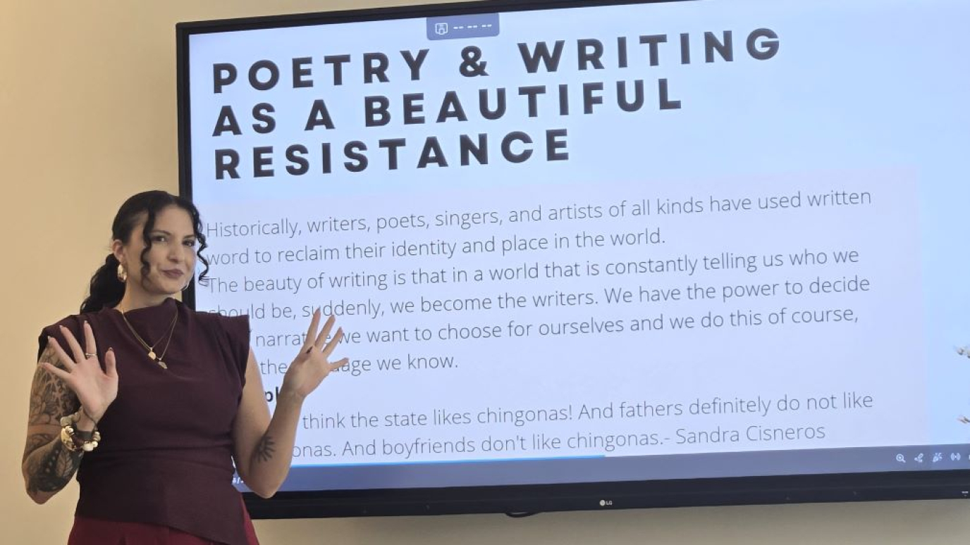 Hispanic woman standing before large screen with text reading Poetry and writing as a Beautiful Resistance
