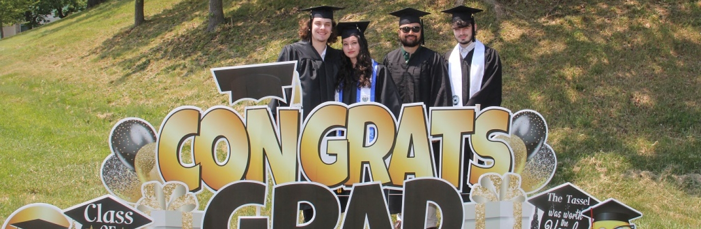 Four graduates in caps and gowns standing among Congrats Grad lawn signs and ornaments