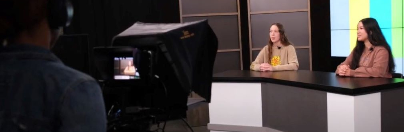Two female students behind a news desk as a third student films them