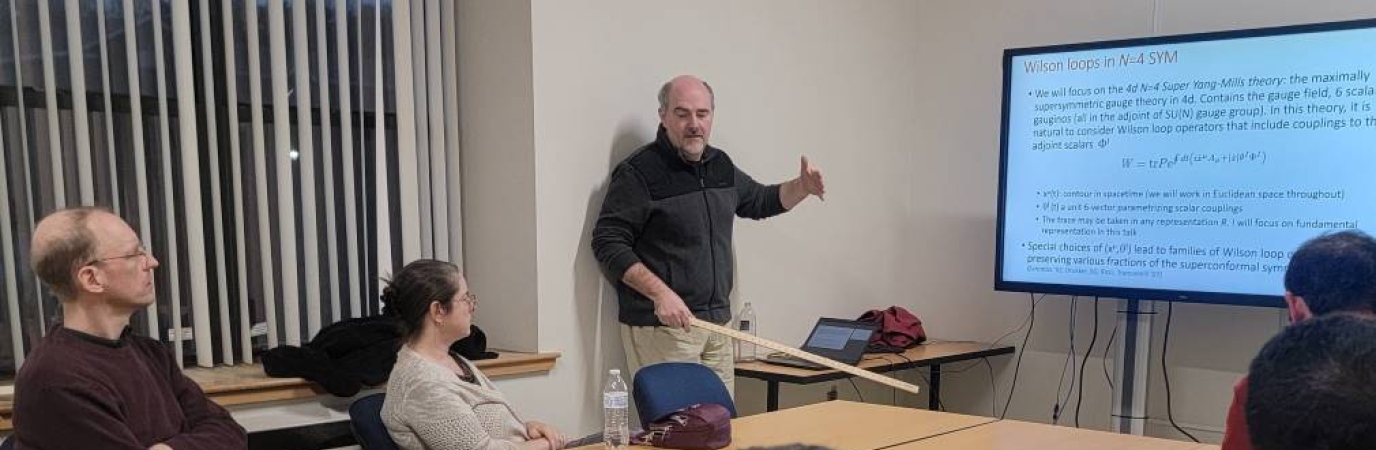 Man holding pointer lectures before and LED screen to a group sitting around a table