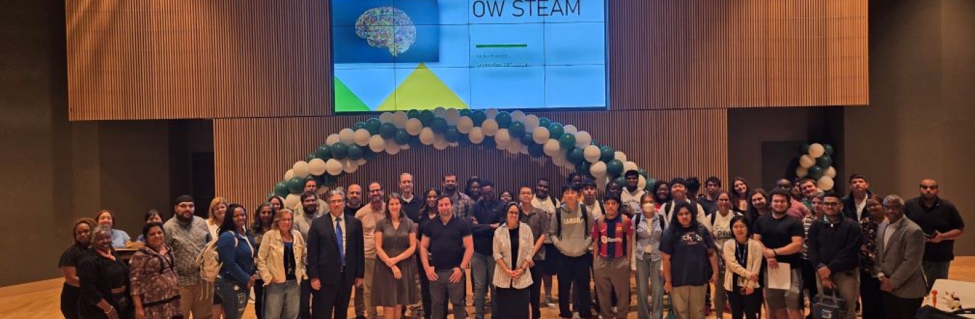 A large group of people from diverse backgrounds pose in an auditorium under a sign that reads OW STEAM