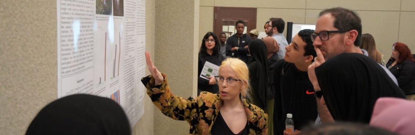 Woman points to scientific poster while speaking while group of seven other people look on