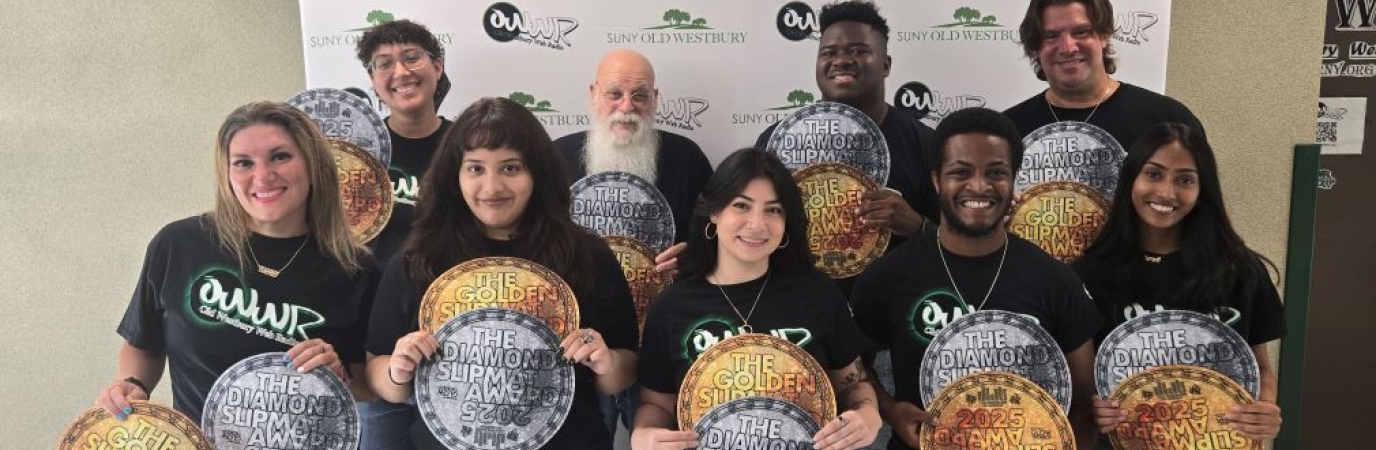 A diverse group of 10 people wearing black tee shirts hold copies of a silver and gold circular award before a step and repeat featuring the college logo and radio station logo