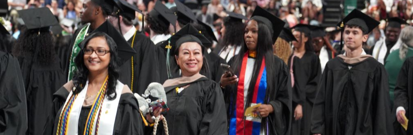 Four students from diverse backgrounds wearing academic regalia march in a procession in a crowded floorspace